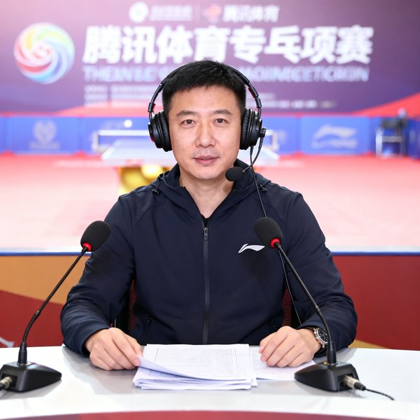 腾讯体育乒乓球专项解说员刘伟国正面照，戴着耳机坐在解说席前，面前摆放着赛事资料和麦克风